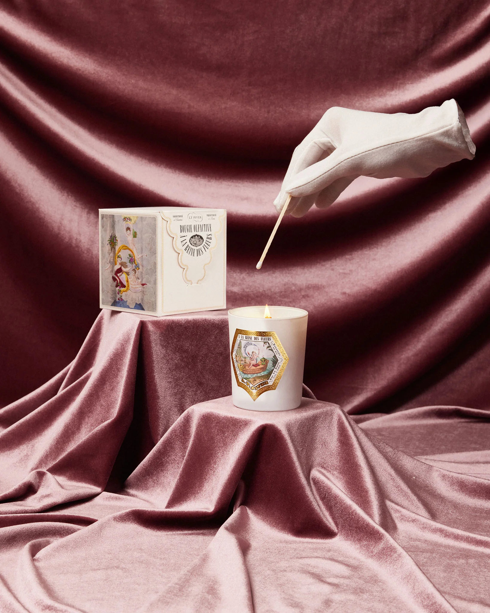 Bougie parfumée L.T. Piver allumée, posée sur un drapé de velours rose, avec son coffret illustré et une main gantée allumant la flamme.