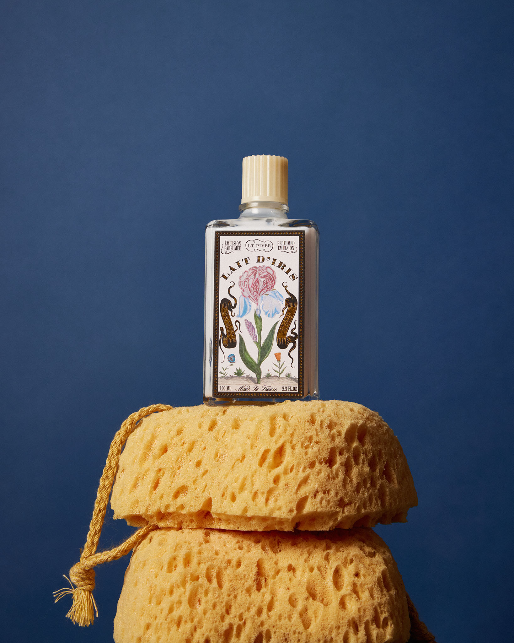Flacon de Lait d’Iris posé sur deux éponges jaunes avec fond bleu, mettant en valeur le design floral du produit.